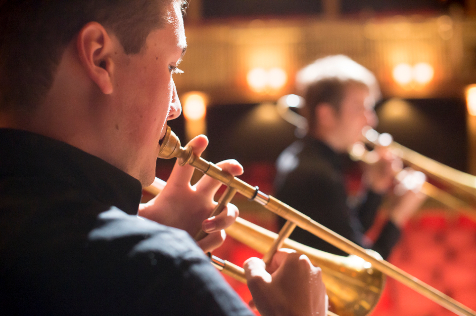 Male student, performing the trombone, with other students performing on the trombone, in a well lit theatre.